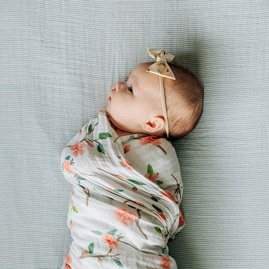 baby laying on a micro grey crib sheet swaddled in a peach blossom blanket