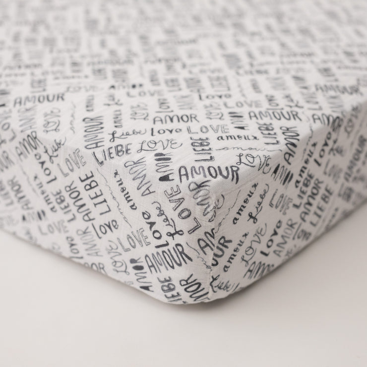 cotton muslin crib sheet with the word "love" in multiple different languages in grey writing on a white background